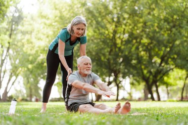 Kıdemli, gri saçlı adam park sporu konseptinde antrenörle esneme egzersizleri yapıyor.