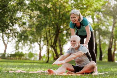 Kıdemli fitness eğitmeni, yaşlı bir adamın dışarıda egzersiz yaparken bacaklarını esnetmesine yardım ediyor. İkisi de gülümsüyor ve birlikte egzersiz yapmanın faydalarından keyif alıyorlar.