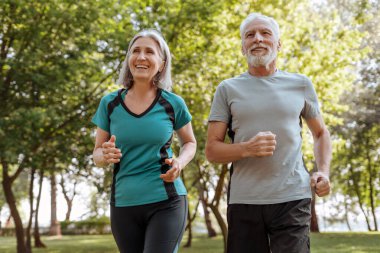 Yaşlı çift parkta mutlu mesut koşuyor, emekliliklerinde aktif yaşam tarzlarını kucaklıyor. Gülümseyen, spor kıyafetler giyen, doğanın tadını çıkaran, canlılığıyla diğerlerine ilham veren. Spor konsepti