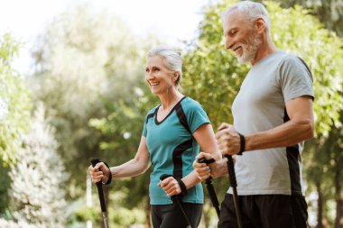 Aktif kıdemli çift yemyeşil bir parkta kuzeyli yürüyüş yaparak aktif ve sağlıklı bir yaşam tarzını kucaklıyor. Spor konsepti