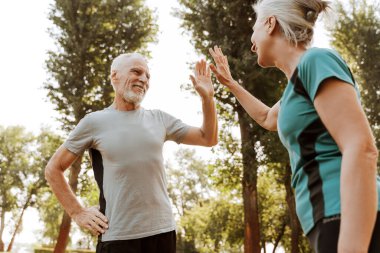 Gri saçlı ve spor giysili bir kadın ve erkek spor kıyafetleri olan bir beşlik çakıyor ve fitness hedeflerine birlikte ulaştıkları için mutlular.