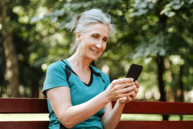 Yaşlı bir kadın parkta mola veriyor, spor yaptıktan sonra akıllı telefona bakıyor, doğanın tadını çıkarıyor, aktif bir yaşam tarzı sergiliyor. Çevrimiçi teknoloji kavramı