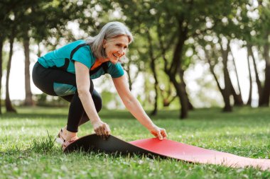 Aktif son sınıf öğrencisi gri saçlı kadın yeşil bir parkta kırmızı yoga minderini açarken gülümsüyor ve açık hava antrenmanına hazırlanıyor.