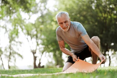 Gülümseyen son sınıf öğrencisi, parkta çimenli bir alanda turuncu bir yoga minderini açıyor. Rahatlatıcı bir açık hava antrenmanına hazırlanıyor. Spor konsepti