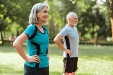 Spor kıyafetleri giyen yaşlı bir adam ve kadın ellerini kalçalarına koyup parkta durup dışarıda egzersiz yapmaya hazırlanıyorlar.