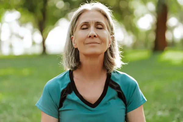 Sağlıklı yaşlı kadın, gözleri kapalı parkta meditasyon yaparken egzersiz yapıyor. Atletik kıyafetler giyiyor ve yüzünde sakin bir ifade var. Denge kavramı