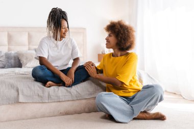 Gülümseyen Afro-Amerikan anne ve kızının portresi lotus pozunda dinleniyor, yatak odasında konuşuyor. Çocukluk, annelik, aile kavramı.