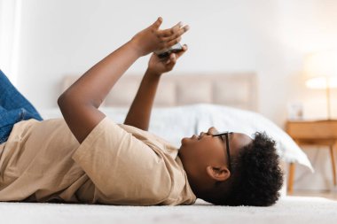 Gözlüklü gülümseyen Afrikalı Amerikalı çocuğun portresi cep telefonu kullanıyor, yatak odasında yerde yatıyor, oyun oynuyor.