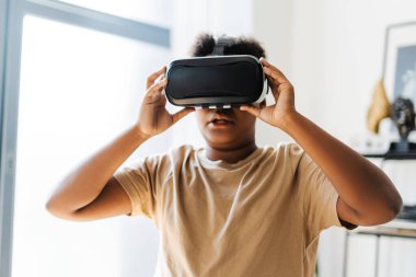 Amazed African American boy using VR headset while playing at futuristic game at home. Modern technology concept clipart