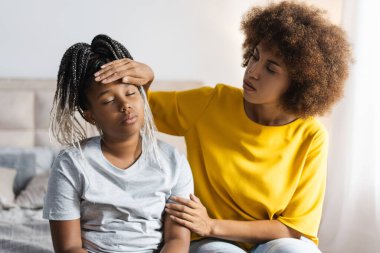 Afrika kökenli Amerikalı annenin portresi evde kızının alnındaki sıcaklığı ölçüyor. Çocukluk, annelik, sağlıklı bakım konsepti