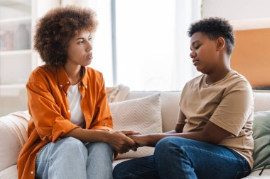 Kıvırcık Afrikalı Amerikalı anne rahat bir dairede genç oğluyla kanepede tartışıyor. Aile sorunları kavramı