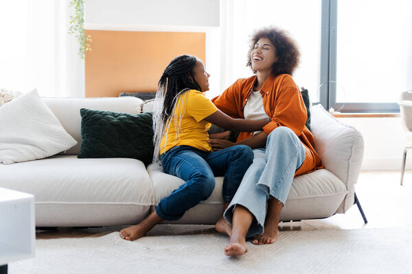 Симпатичная американка из Африки щекочет мать на диване дома. Забавная семейная женщина и ребенок веселятся вместе