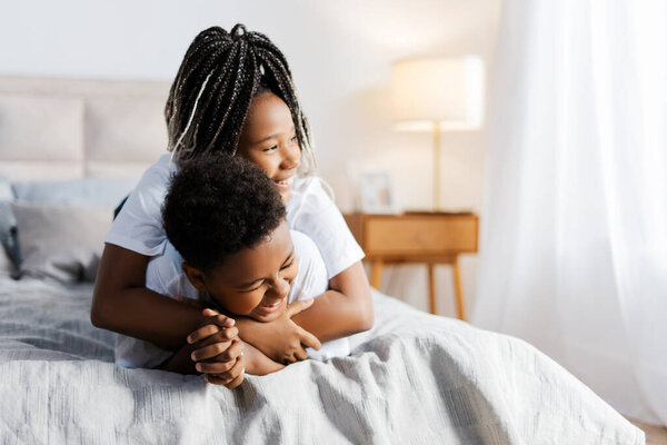 Smiling African American siblings hugging while resting on the bed at home. Childhood, relatives concept