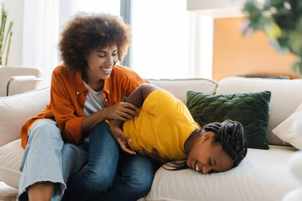 Funny African American mother tickling her child girl, sitting on sofa at home. Concept of childhood, motherhood, family, fun