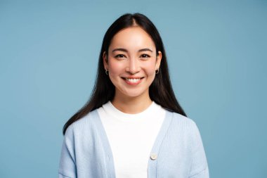 Mavi kazak giyen genç, gülümseyen Asyalı kadın mavi arka planda tek başına kameraya bakıyor. Reklam kavramı