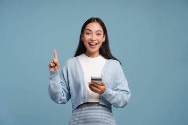 Heyecanlı bir kadın elinde akıllı telefonuyla parmaklarıyla işaret ederek mavi arka planda tek başına duran kameraya bakıyor.