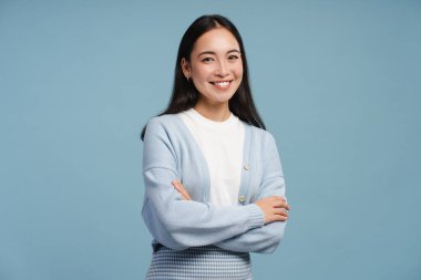 Gülümseyen, kollarını kavuşturmuş genç Asyalı bir kadın, kameraya bakıyor, mavi arka planda tek başına duruyor, stüdyoda poz veriyor. Reklam kavramı