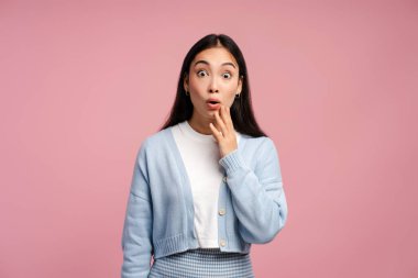 Uzun siyah saçlı, şaşırmış genç bir kadın pembe arka planda poz verirken eliyle ağzını kapatıyor.