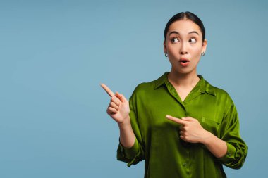 Şaşırmış Asyalı iş kadını mavi arka planda dururken iki parmağını yana gösteriyor. Reklam kavramı