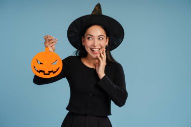 Cadı kostümü giyen heyecanlı genç Asyalı kadın mavi arka planda Cadılar Bayramı balkabağı süslemesi tutuyor. Kutlama konsepti