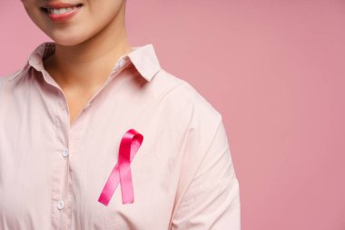 Pembe gömlekli, ipek kurdeleli, pembe arka planda poz veren, göğüs kanseri farkındalığı olan gülümseyen bir kadın görüntüsü.