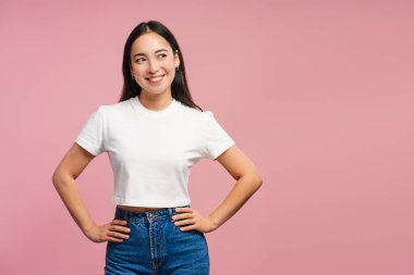 Çekici, gülümseyen Asyalı kadın ellerini pembe arka plan fotokopi alanındaki kalçalarına koyup poz veriyor. Stüdyoda poz veren güzel bir model. Alışveriş konsepti