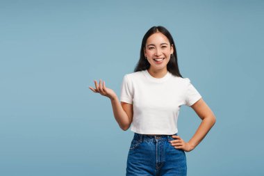 Gülümseyen, çekici Asyalı kadın elinde bir şey olan, mavi arka planda izole edilmiş bir şeyi açıklayan, kopya alan. Stüdyoda poz veren gündelik kıyafetler giyen güzel bir manken. Reklam kavramı