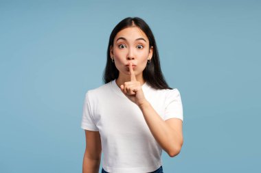 Heyecanlı Asyalı kadın, parmakları dudaklarında, mavi arka planda izole edilmiş kameraya bakarak sessiz jest yapıyor. Gizli konsept