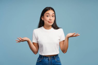 Confused young Asian woman shrugging with raised arms and open palms standing isolated on blue background. Advertisement concept clipart