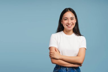 Kollarını kavuşturmuş gülümseyen Asyalı kadın mavi arka plandaki kameraya bakıyor, kopyalama alanı. Reklam kavramı