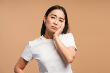 Güçlü bir diş ağrısı çeken genç Asyalı kadın yanağa dokunuyor, bej arka planda izole edilmiş kameraya bakıyor. Sağlık hizmeti kavramı