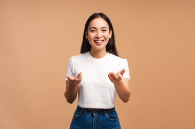 Stüdyo portresi, uzun siyah saçlı, beyaz tişört ve kot giyen, elleri bej arka planda uzanmış, gülümseyen ve el kol hareketi yapan genç bir kadın.