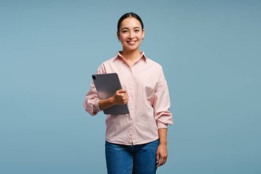 Gülümseyen genç bir kadın elinde tabletle mavi bir arka plana karşı duruyor, teknoloji meraklısı, modern bir havayı somutlaştırıyor. Dijital göçebe yaşam tarzını, pozitifliği ve bağlantıyı temsil ediyor.