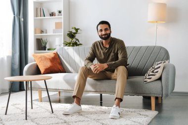 Orta yaşlı yakışıklı Hintli adam evdeki modern apartman dairesinde kanepede oturmuş kameraya bakarak gülümsüyor. Uzak görev konsepti