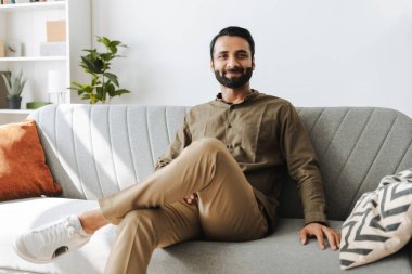 Kendine güvenen girişimci, evdeki koltukta oturan gülümseyen Hintli adam oturma odasındaki kameraya bakıyor. İç mekan kavramı