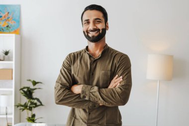 Kendine güvenen işadamı, ofiste kollarını kavuşturup gülümseyen yakışıklı Hintli adam. Başarılı iş konsepti