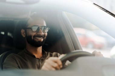 Gülümseyen orta yaşlı Hintli işadamı bir arabanın sürücü koltuğunda oturuyor, direksiyonu tutuyor ve gülümsüyor. Güneş gözlüğü ve yakalı bir gömlek giyiyor.