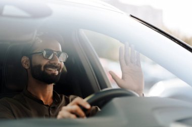 Genç işadamı güneşli bir günde arabasını sürerken mutlu bir şekilde el sallıyor, şehir ortamında güven ve neşe saçıyor, modern bir yaşam tarzını somutlaştırıyor.