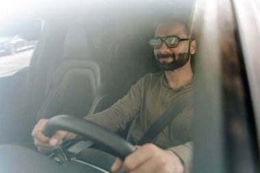 Güneşli bir günde kendine güvenen bir işadamı araba kullanıyor. Rahat ve mutlu görünüyorsun. Güneş gözlüğü takıyordu. Lüks ve rahat bir modern iç mekanda yolculuğun tadını çıkarıyorum.