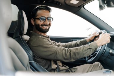 Neşeli Hintli işadamı güneşli bir günde lüks bir araba kullanıyor, gülümsüyor ve güneş gözlüğü takıyor, bir yolculukta büyük bir sevinç ve başarı yaşıyor.