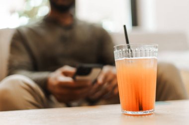 Bir bardak buzlu meyve suyu ve siyah bir pipet, akıllı telefonunu kullanarak bir kanepede dinlenen bir adamın önündeki ahşap masada oturuyor.