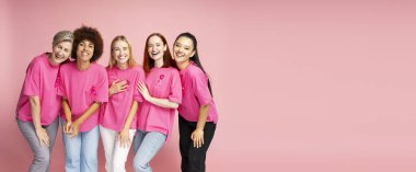 Farklı yaş ve etnik kökenlere sahip beş farklı kadın gülüyor ve pembe kurdeleli pembe gömlekler giyip birlikte poz veriyorlar.