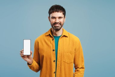 Gülümseyen, çekici Latin adam şık sarı gömlek giyiyor, cep telefonunu boş beyaz ekran ile tutuyor, mavi arka planda izole edilmiş kameraya bakıyor, reklam, çevrimiçi teknoloji konsepti