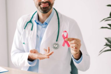 Beyaz önlüklü ve şık gözlükler takan profesyonel bir doktor pembe kurdele tutuyor, kameraya bakıyor ve seçici bir şekilde odaklanıyor. Göğüs kanseri farkındalığı konsepti