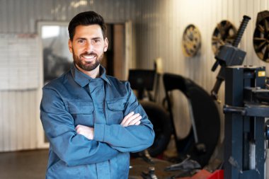 Profesyonel tulumlu kendine güvenen tamirci, araç gereç ve lastiklerle çevrili yoğun atölyede gülümsüyor, araba tamir sektöründe uzmanlık ve tutku sergiliyor