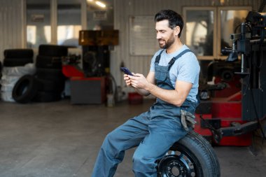 Tamirci araba tamirhanesinde oturuyor, telefonunu kontrol ederken gülümsüyor, mevsimlik lastik değiştirmeye ara veriyor.