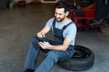 Mekanik bir atölyede lastik üzerinde oturmuş gülümseyen bir tamirci, işe ara veriyor ve akıllı telefonuna göz atıyor.