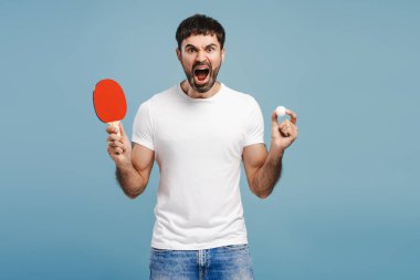 Heyecanlı, mutlu, şık giyinen Latin bir adam tenis raketi tutuyor ve haykırıyor mavi arka planda izole edilmiş kameraya bakıyor. Zafer konsepti