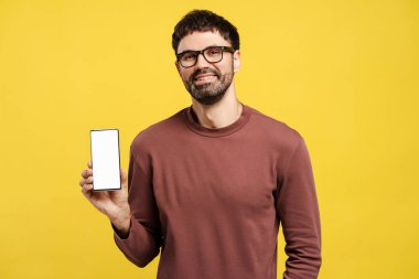 Yakışıklı, çekici, sakallı, gözlüklü, cep telefonu takan, boş, beyaz ekranlı, sarı arka planda izole edilmiş bir model. Reklam kavramı