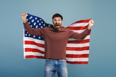 Aşırı neşeli, sakallı bir adam çığlık atıyor, elinde Amerikan bayrağı, kameraya bakıyor, mavi arka planda tek başına duruyor. Özgürlük, bağımsızlık günü, ABD konsepti oylaması